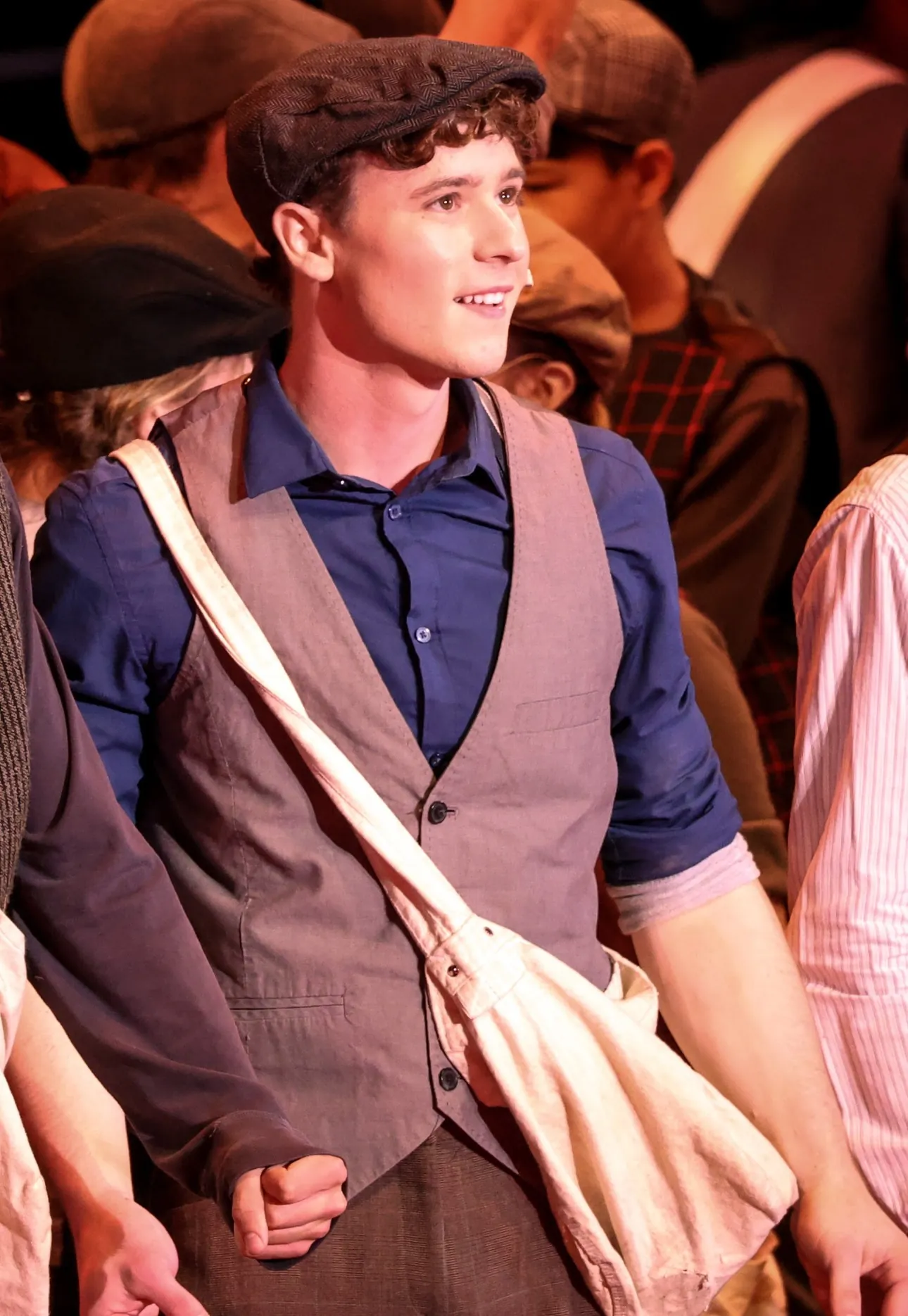 Caleb Mackie on stage in costume, wearing a newsboy cap and vest